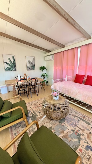 A room with a bed, a dining table with chairs, a round wicker coffee table, and a patterned rug. - Oceanfront 2BR/2BA Home · 2 Kings, Fast WiFi, AC, BBQ & Fire Pit (Ewa Beach)