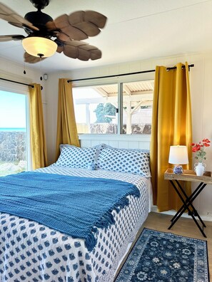 A bedroom with a bed, a ceiling fan, a window with curtains, a bedside table with a lamp, and a rug. - Oceanfront 2BR/2BA Home · 2 Kings, Fast WiFi, AC, BBQ & Fire Pit (Ewa Beach)