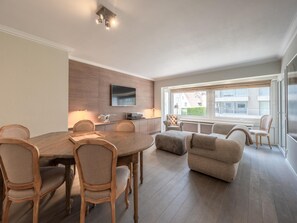 A modern living room with a dining area, a flat-screen TV mounted on the wall, and a large window with a view of buildings. - Apartment in Knokke Near Albert Beach (Knokke-Heist)