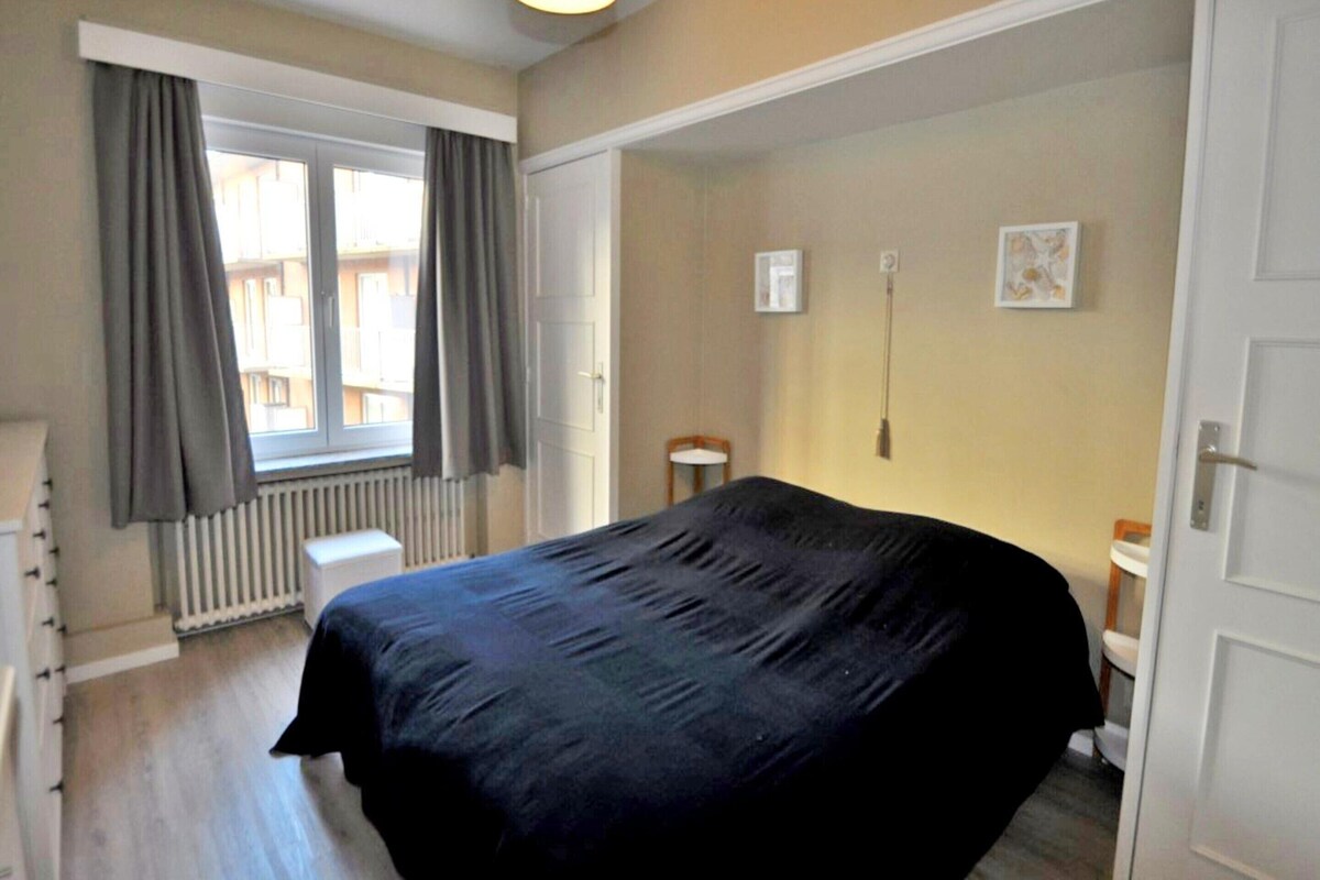A bedroom with a bed, a radiator, a window with curtains, a bedside table, and a door.