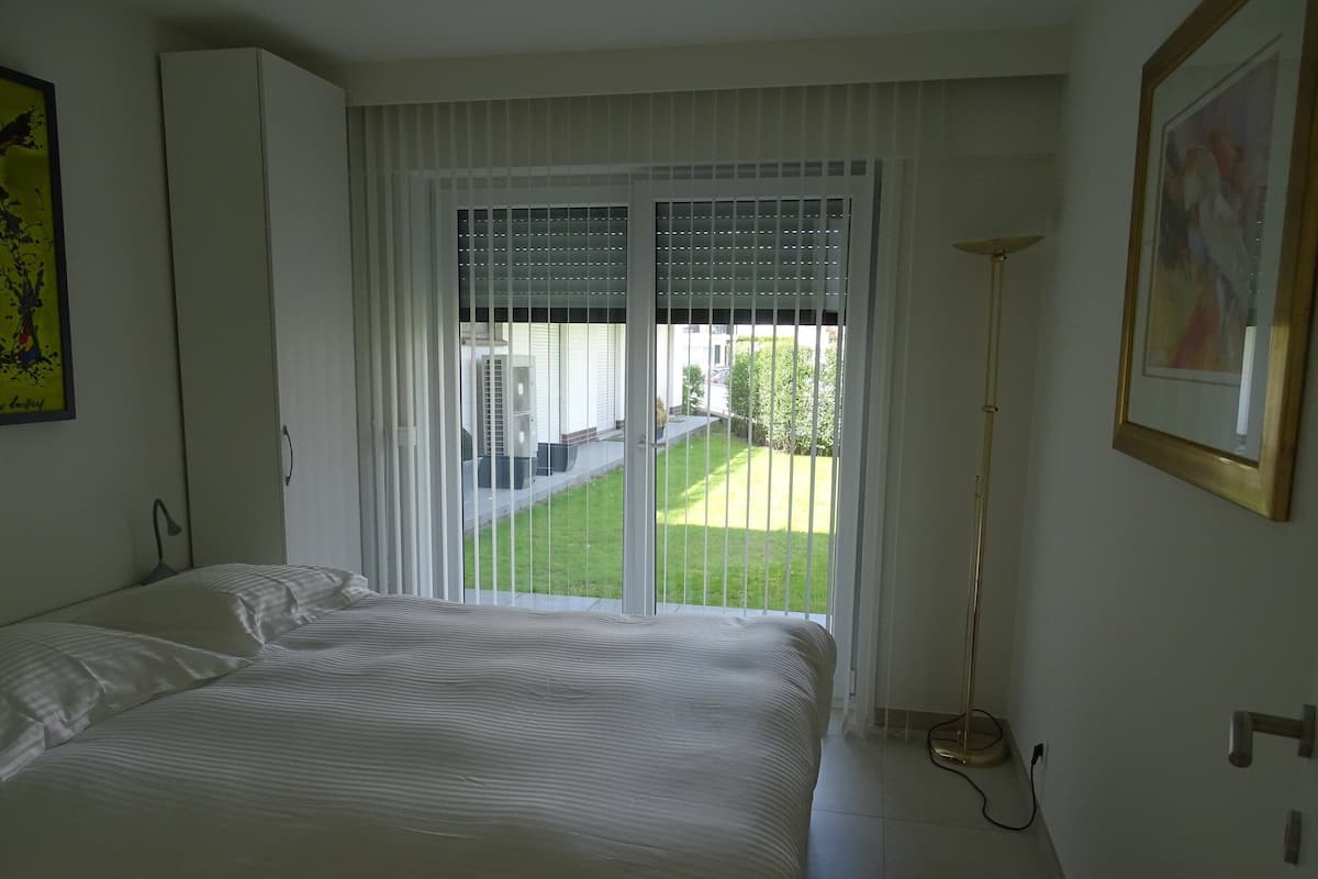Ein Schlafzimmer mit einem Bett, einem Fenster mit Jalousien, einer Stehlampe und einem Bild an der Wand.