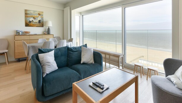A modern living room with a sofa, coffee table, and dining area. A large window offers a view of the beach. - Cozy Knokke Beach Escape (Knokke-Heist)