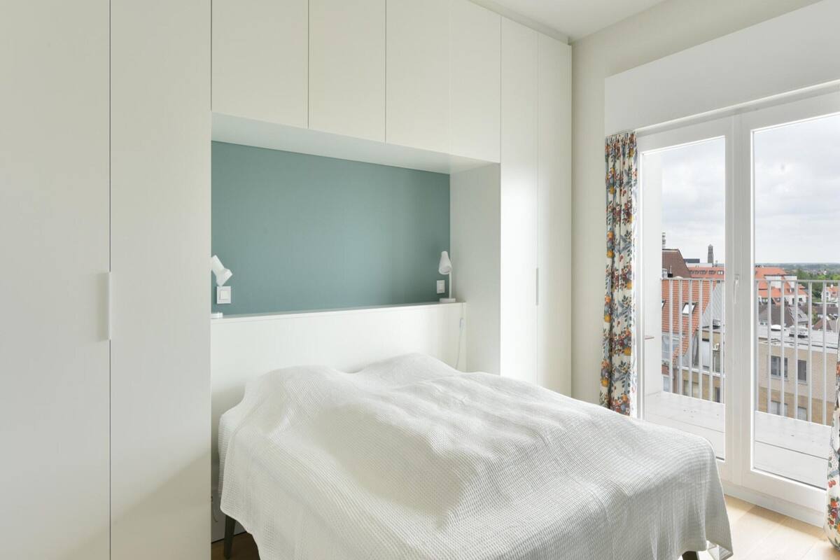 A bedroom with a bed, a white wardrobe, a small shelf, and a window with a view of buildings.