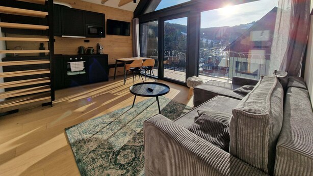 A modern interior with a kitchen, dining area, and a large window offering a view of a town. - Apartment in Krkonose With Mountain Views (Pec pod Snezkou)