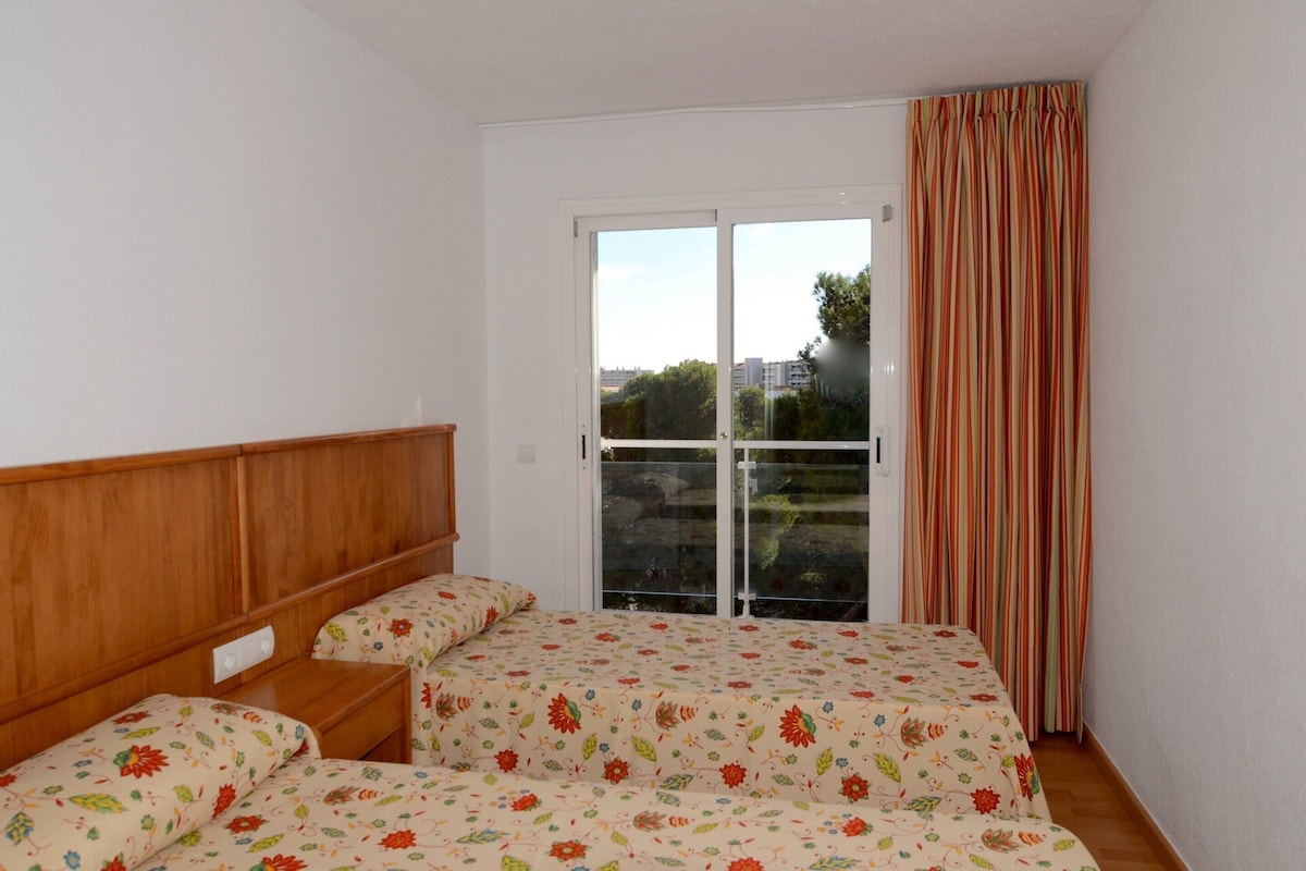 A bedroom with a wooden headboard, a bed with floral bedding, a window with curtains, and a view of buildings outside.