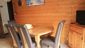Ein Zimmer mit Holzwänden, einem runden Holztisch, vier Stühlen, einem Fernseher und einer Wanduhr.
