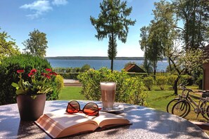 An open book, a pair of sunglasses, and a glass of beverage on a table with a view of a lake and greenery. - Seepark Heidenholz, Plau am See (Plau am See)