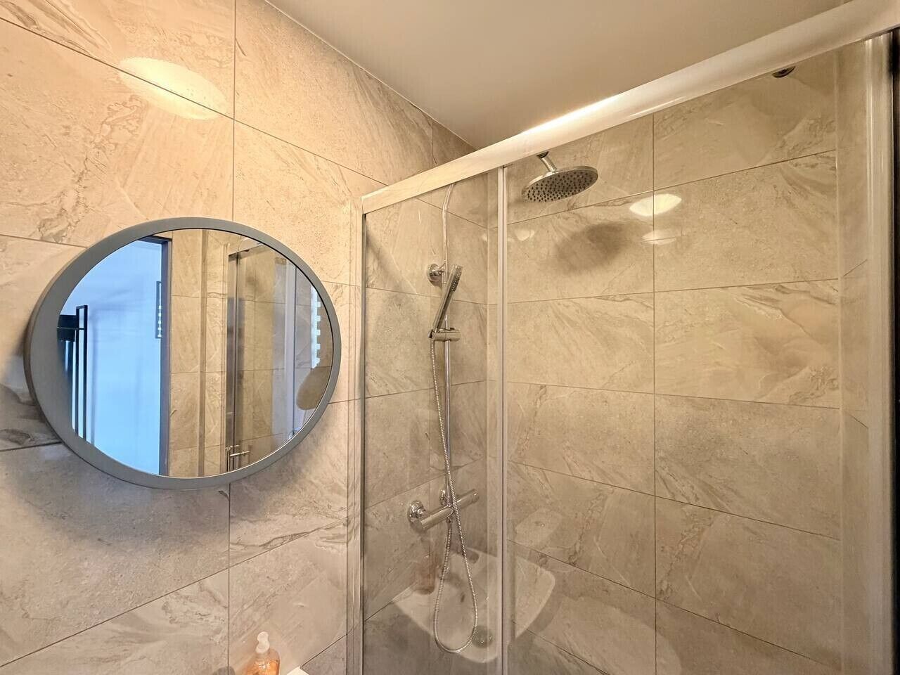 A modern bathroom with a glass-enclosed shower, a round mirror, and marble tiles.