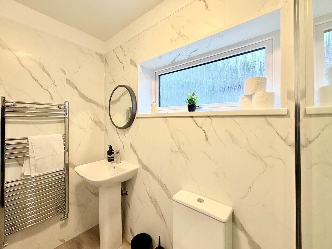 A bathroom with a marble wall, a sink, a toilet, a towel rack, and a window with a plant.