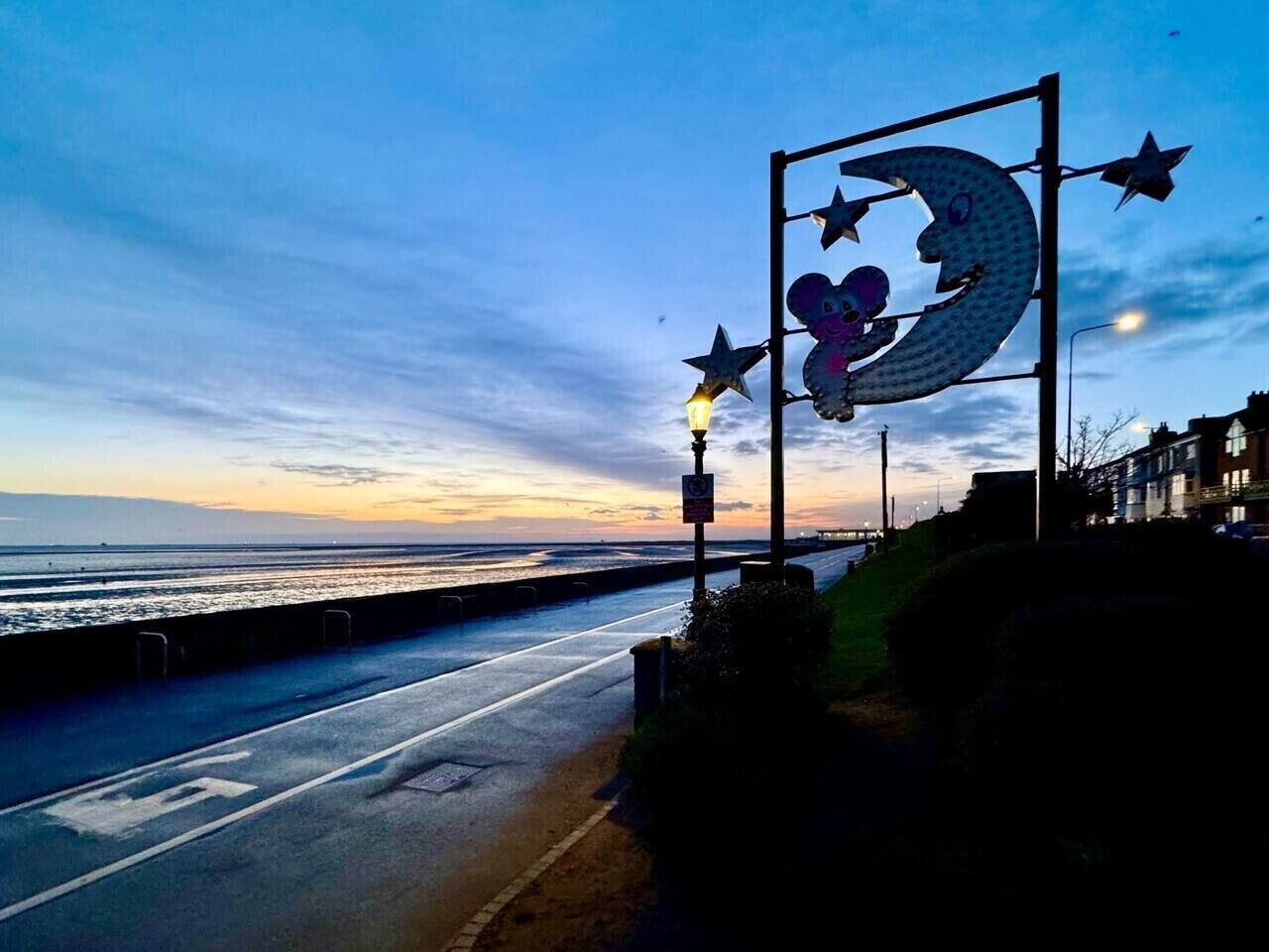 Una carretera costera con una acera peatonal, una farola y un letrero de metal decorativo con estrellas y una luna creciente.