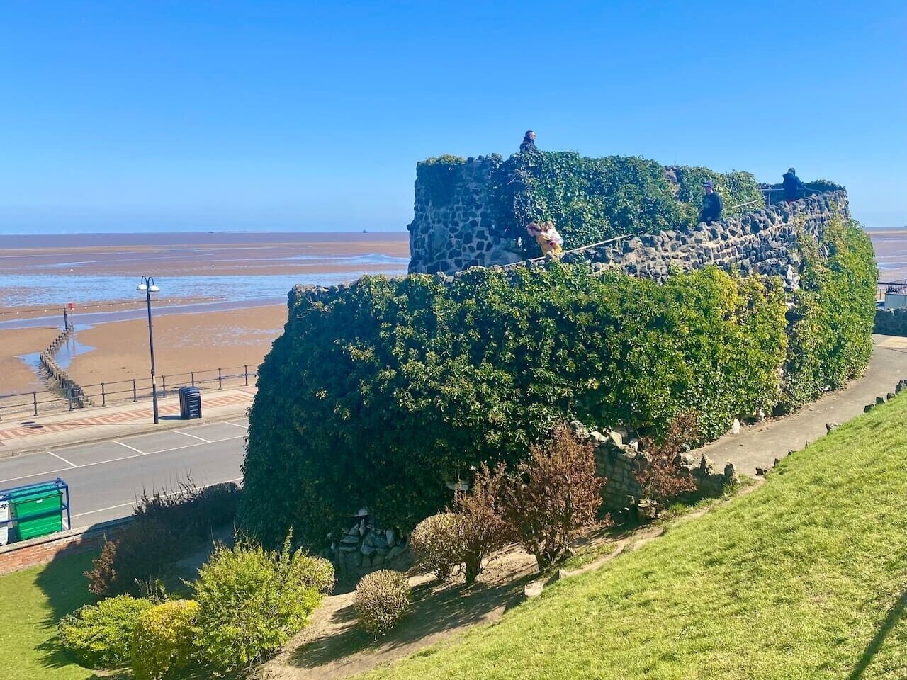 A coastal fortification with a grassy foreground, a paved road, and a clear view of the beach and ocean.