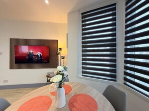 A modern living room with a flat-screen TV mounted on the wall, a dining table with a vase of flowers, and large windows with blinds. - 5 Spacious 2-Bed Flat Sleeps 4 Secure Parking WiFi (Cleethorpes)