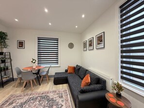 Un salon moderne avec un canapé d’angle, une table à manger avec des chaises, un petit tapis et une plante en pot.