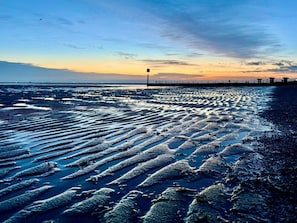 Une plage avec des motifs dessinés sur le sable humide, une jetée en arrière-plan et un ciel dégagé.