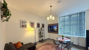 Ein modernes Wohnzimmer mit einer Couch, einem Esstisch mit Stühlen, einem an der Wand montierten Fernseher und einem großen Fenster mit Jalousien.