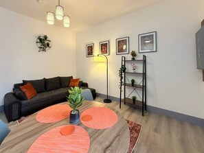 A modern living room with a sofa, a coffee table with orange accents, a floor lamp, and a shelving unit with plants. - 3 Spacious 1-Bed Flat Sleeps 3 Secure Parking WiFi (Cleethorpes)