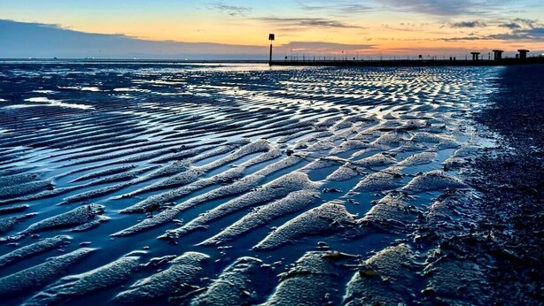 Ein Strand mit feuchten Sandmustern, ein Steg im Hintergrund und ein klarer Himmel.