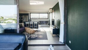 A modern interior with a sofa, a wicker chair, a dining area with stools, a kitchen, and a sliding glass door leading to a balcony. - Jan Sofat LUX 2BR Penthouse Pool + Mountain View (Willemstad)