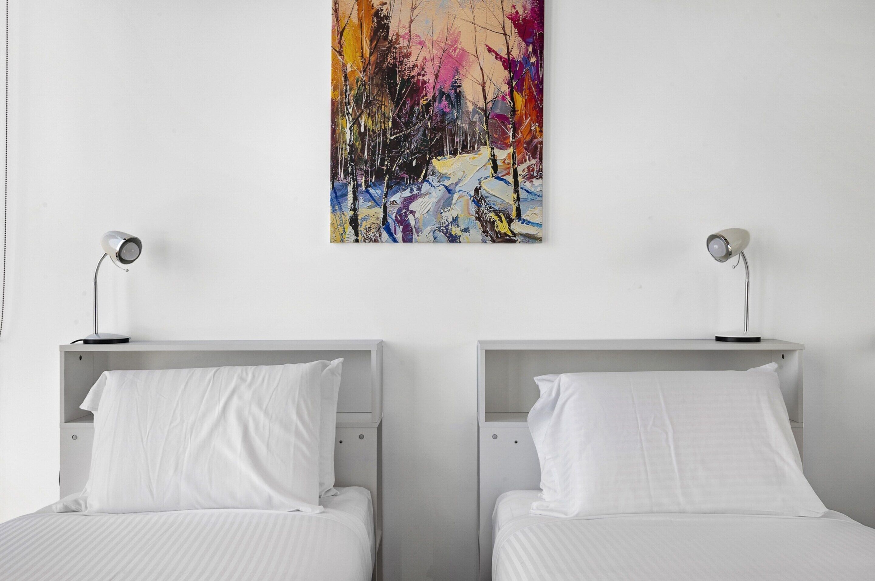 Ein Hotelzimmer mit zwei Einzelbetten, einer weißen Wand und einem farbenfrohen abstrakten Gemälde darüber.