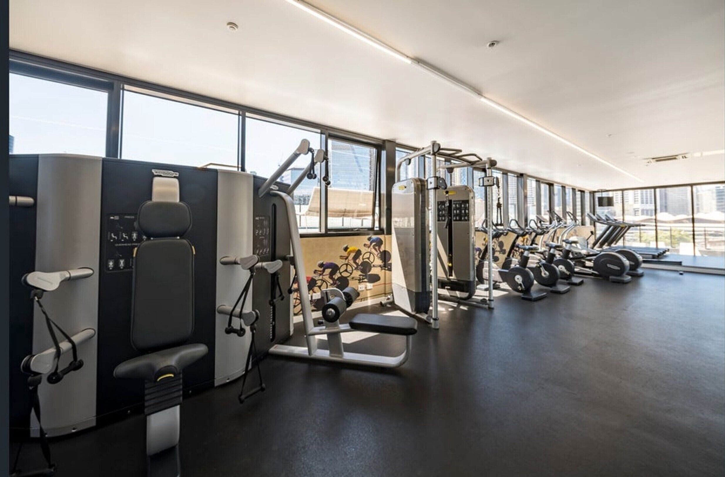 A gym with various exercise machines, large windows, and a view of buildings.