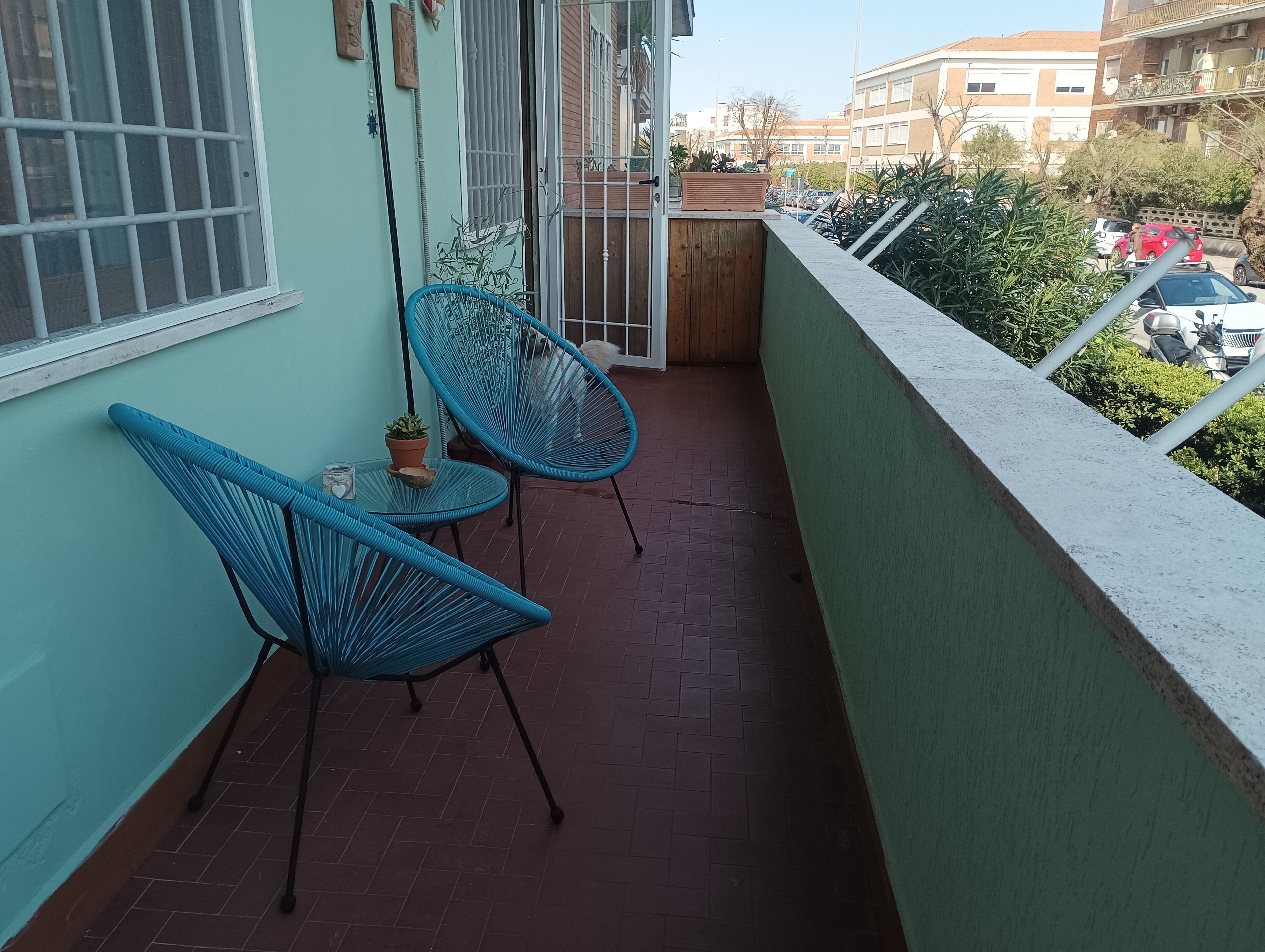 A balcony with two blue chairs, a small table with a potted plant, and a view of parked cars and buildings.