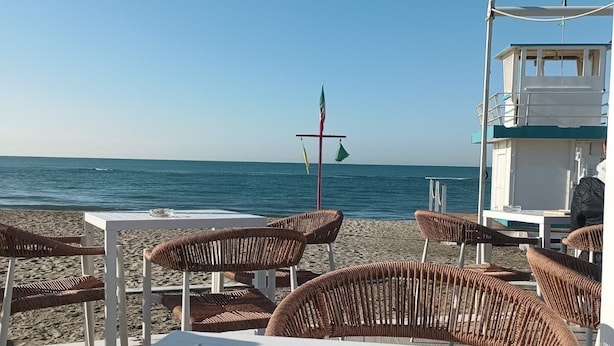 A beachside café with wicker chairs and tables, a view of the sea, and a lifebuoy on a pole. - Domus Quinqueremi (roma)