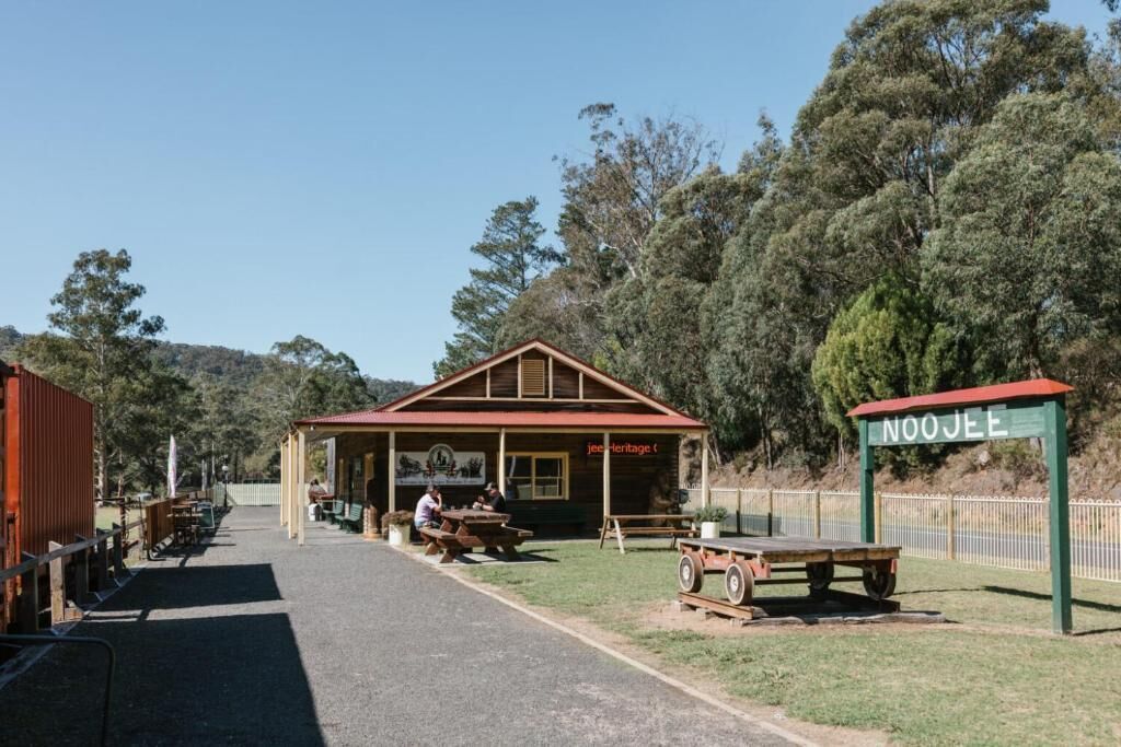 A wooden building with a red roof, labeled 'Noojee Heritage,' with picnic tables and a signboard.