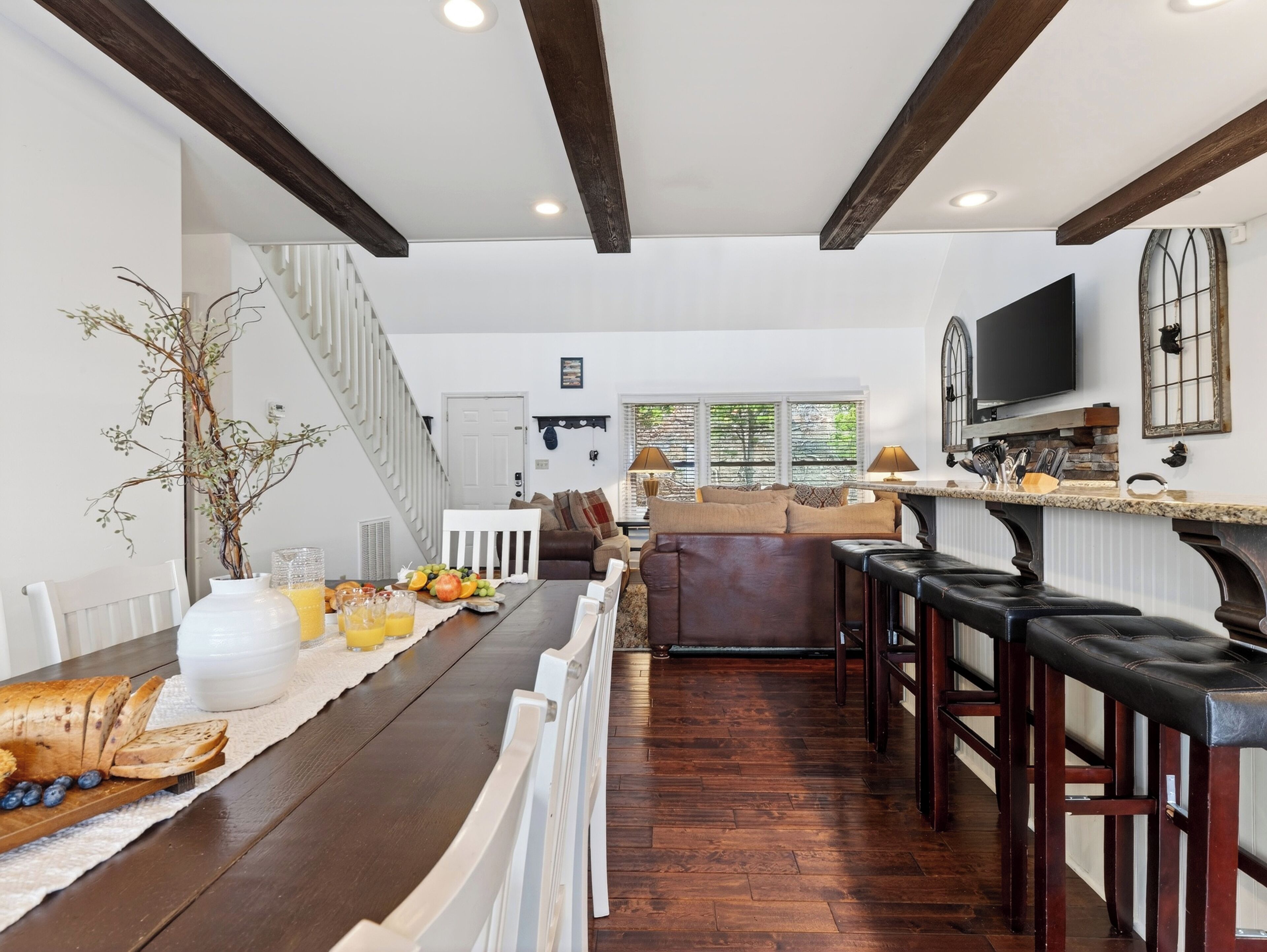 A dining area with a long wooden table, white chairs, and a white railing. There is a staircase leading to another room, a television mounted on the wall, and a window with a view of trees.