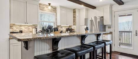 Una cocina con armarios blancos, encimera de granito, refrigerador de acero inoxidable y una zona de bar con taburetes.