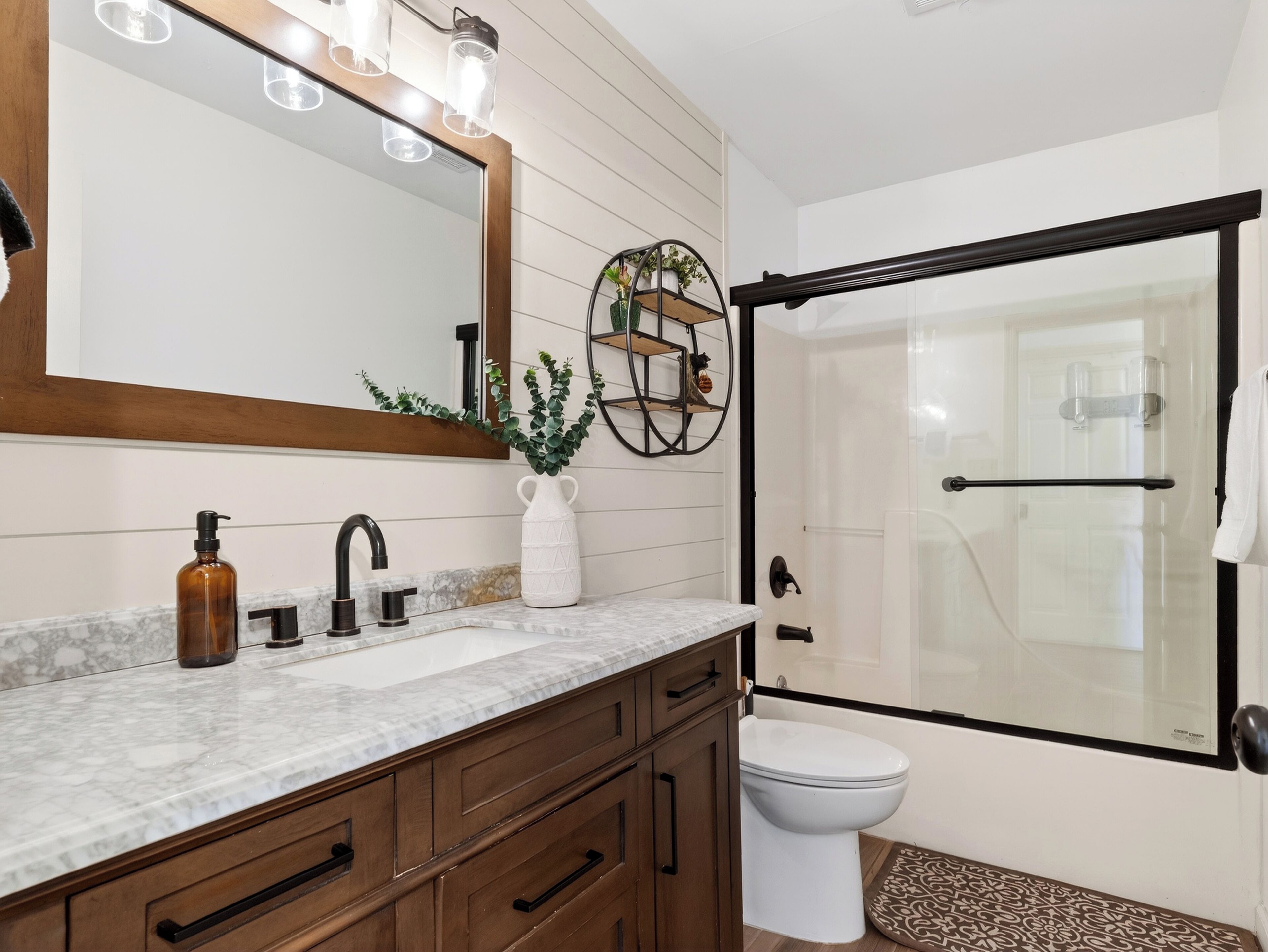 A bathroom with a marble countertop, wooden cabinets, a white toilet, a glass shower enclosure, and a mirror with lights.