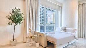 Une chambre d’hôtel moderne avec un grand lit, une plante en pot, une petite table ronde et une vue sur le paysage urbain grâce à de grandes fenêtres.
