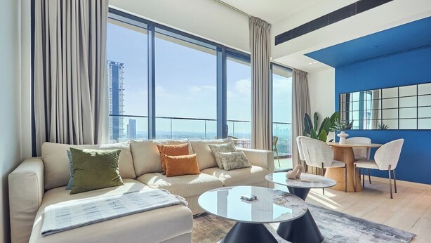 A modern living room with a sofa, coffee table, and chairs, large windows with curtains, and a view of the cityscape. - Binghatti Corner - 1508 (Dubai)