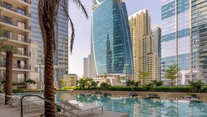 A modern hotel with a pool area, palm trees, and high-rise buildings in the background. - Zada Tower | 1BR, Big Balcony, Bright & Breezy (Dubai)