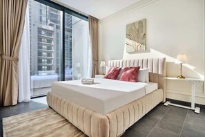 Ein modernes Hotelzimmer mit einem großen Bett, Blick auf die Stadt durch raumhohe Fenster, einem Nachttisch mit Lampe und einem an der Wand hängenden Kunstwerk.