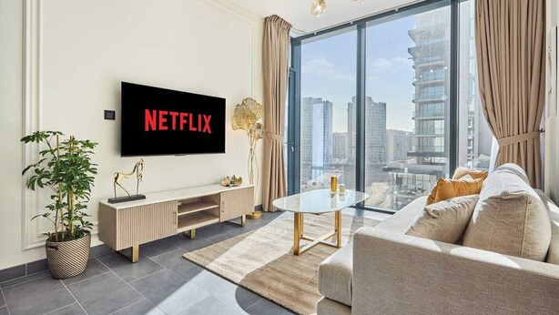 A modern living room with a flat-screen TV, a sofa, a coffee table, a potted plant, and a large window with a city view. - The Crest C Luxury Retreat | Stylish & Serene (Dubai)