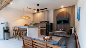 A modern kitchen with a central island, a dining area with wooden chairs, a sofa with cushions, a ceiling fan, and a flat-screen TV mounted on the wall. - Perenenan Home (Pererenan)