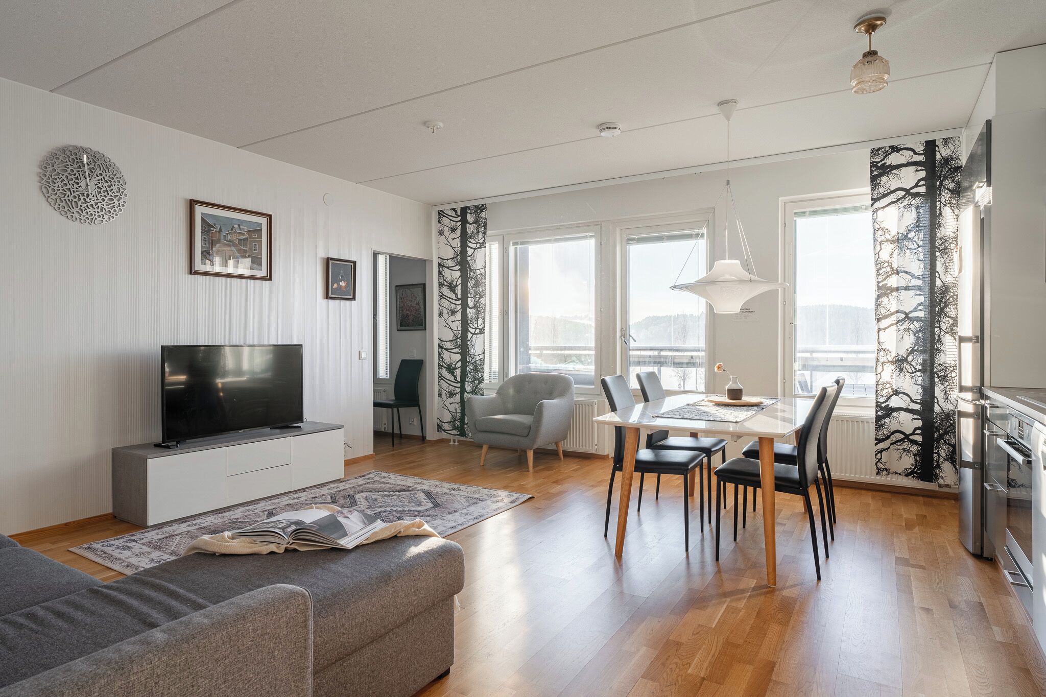 Una sala de estar moderna con un televisor de pantalla plana, un sofá gris, una zona de comedor con una mesa y sillas, y una cocina con estufa.