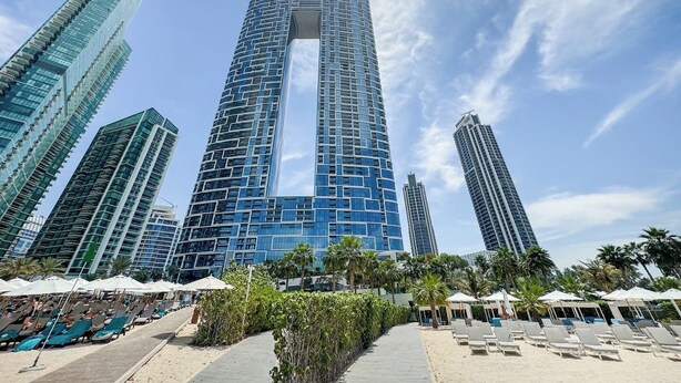 Een modern hoog hotel met een opvallend centraal atrium, omgeven door palmbomen en een zandstrand met ligbedden.