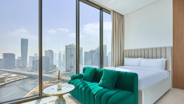 A modern hotel room with a large window, a green sofa, a bed, and a small round table. - Modern Chic Studio Featuring Skyline Views (Dubai)