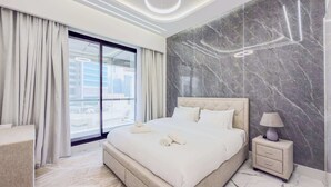 A modern hotel room with a large bed, a nightstand, a lamp, and a view of the city through floor-to-ceiling windows. - Marina View, Fully Furnished, Close to Marina Walk (Dubai)