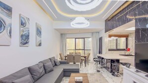 A modern living room with a sofa, dining table, and chairs, a kitchen area with a TV, and large windows offering a city view. - Marina View, Fully Furnished, Close to Marina Walk (Dubai)