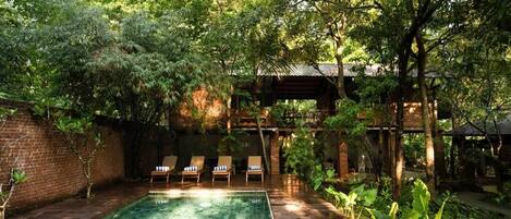 A swimming pool with tiled flooring, surrounded by lush greenery and a brick wall.