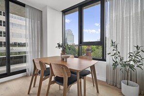 A dining area with a wooden table, four chairs, a vase with a plant, and a potted plant. - Brand New Apartment in Albert Park With Pool & Gym (Melbourne)