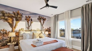 A hotel room with a bed, a ceiling fan, a nightstand, a lamp, a window with curtains, and a desert-themed mural on the wall. - 2 Homes | Hot Tub + Karaoke Lounge + Rooftop + Skyline Views | Min to Broadway (Nashville)
