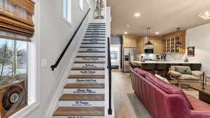 A staircase with steps labeled with names of national parks and monuments, leading to a modern kitchen with wooden cabinets and a red leather bench. - 2 Homes | Hot Tub + Karaoke Lounge + Rooftop + Skyline Views | Min to Broadway (Nashville)