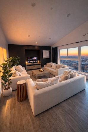 A modern living room with a large sofa, a coffee table, and a TV. There are potted plants and a view of the city through large windows. - Skyline Hilltop 6BR Villa | Best Views in SD (Spring Valley)