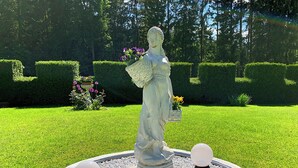 En statue i en rund stensokkel med en have i baggrunden.