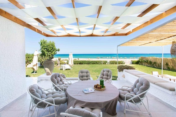 A covered outdoor seating area with a view of the sea, a round table, and cushioned chairs.
