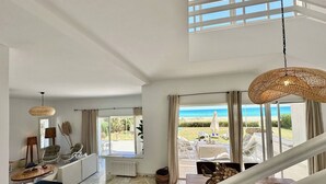 Amplia sala de estar con vista a la playa, zona de comedor con asientos exteriores y una escalera que conduce a un balcón.