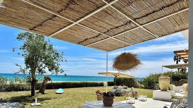Un patio exterior con vista al océano, cubierto por un techo de paja y amueblado con sillas y mesas blancas.